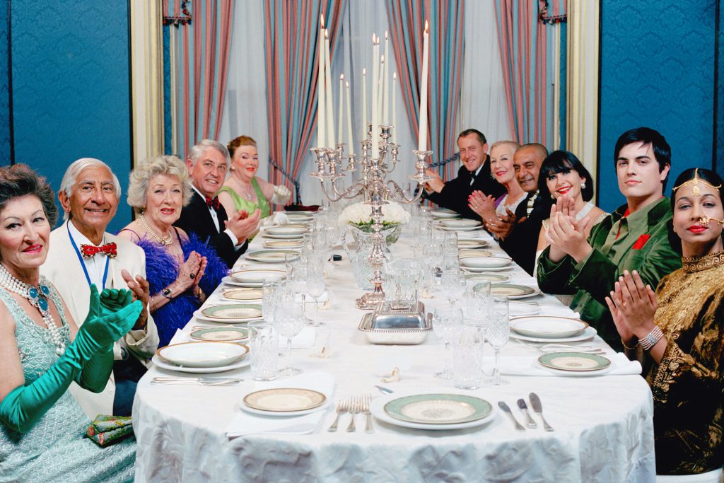 A long dining table set for a formal dinner with 10 formally dressed seated guests applauding the chef.