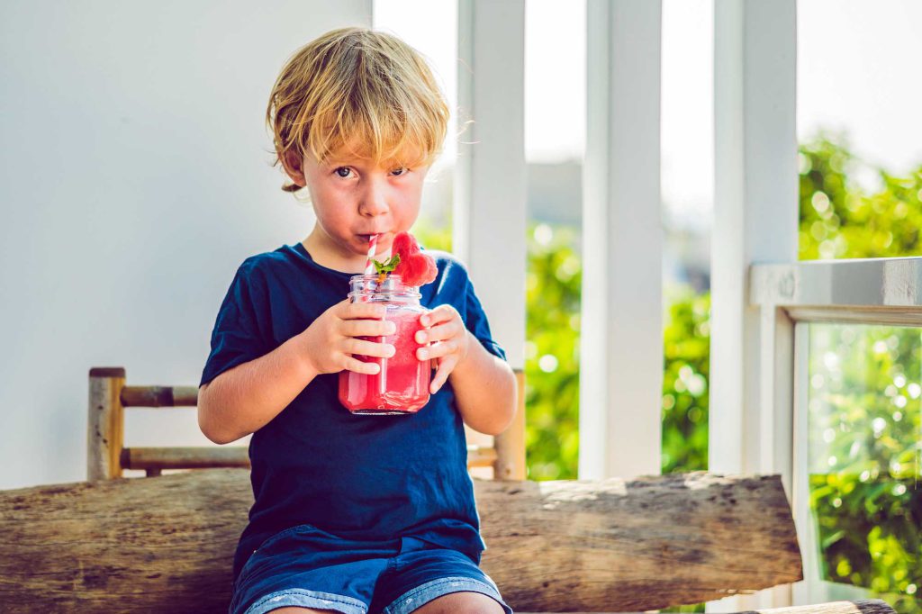 A little boy sits on a kitchen table drinking punch from a mason jar.