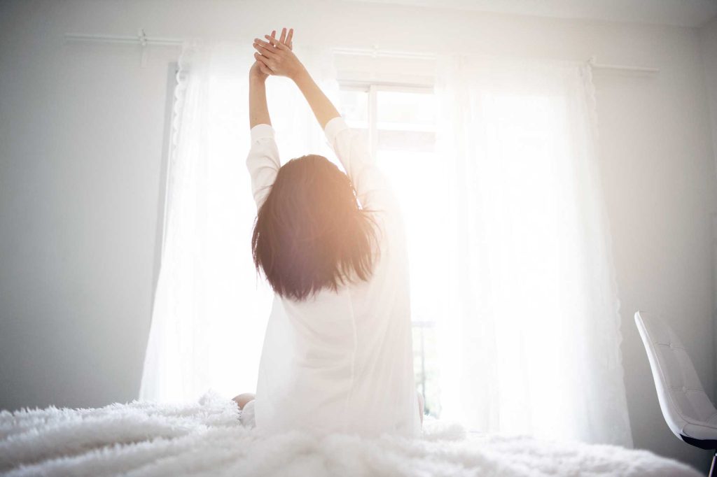 We see a white room and a the back of a woman in a white gown as she sits on her bed and stretches her arms while looking out a window at the sunrise.