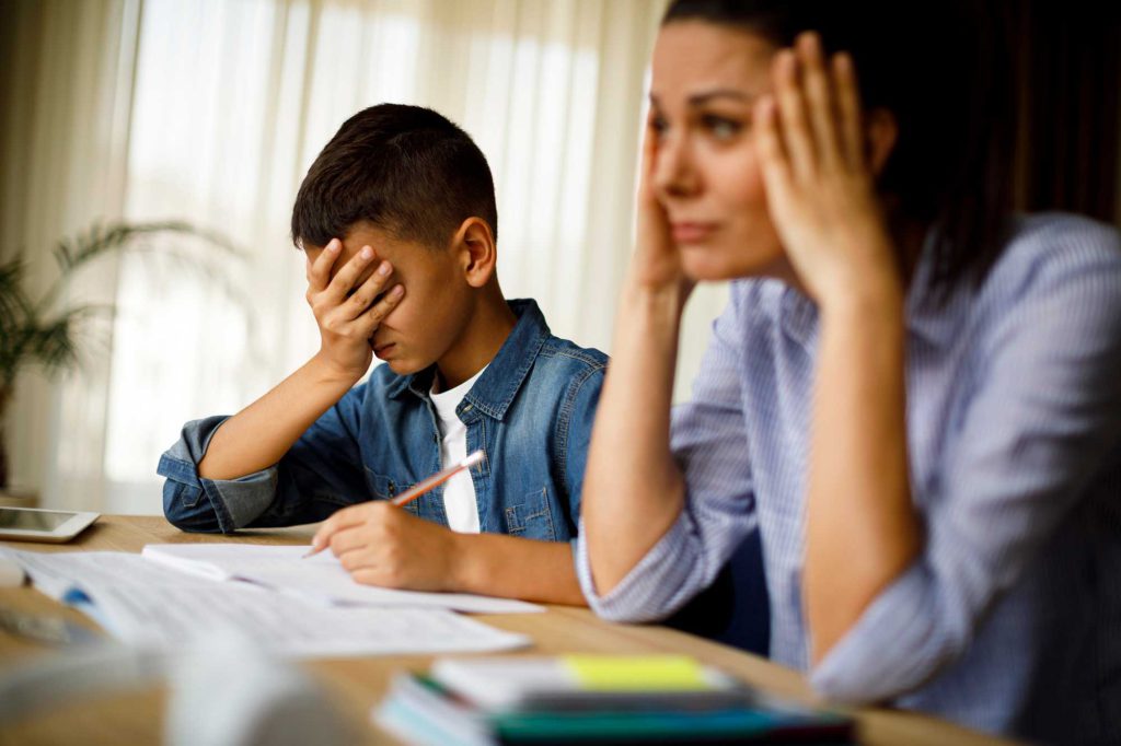 A Hispanic mom shows frustration at helping with homework while her grade school son hides his face.