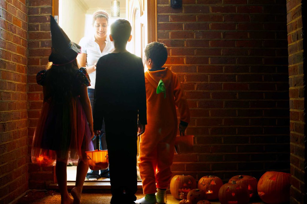 Three kids in costumes are at a front door waiting for the homeowner to hand out candy.