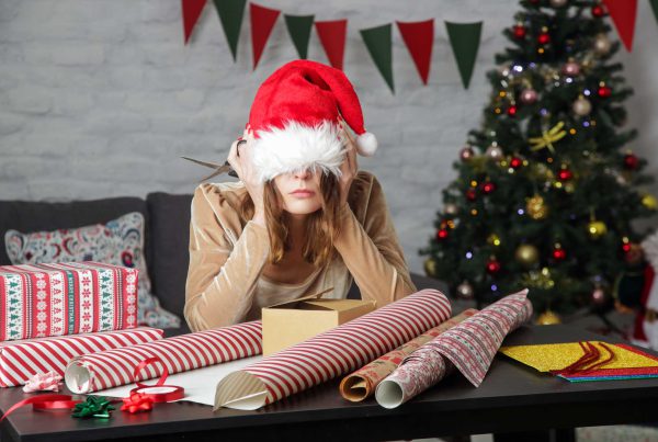 A woman with a Santa hat over her eyes sulks over a table filled with gift wrapping supplies.