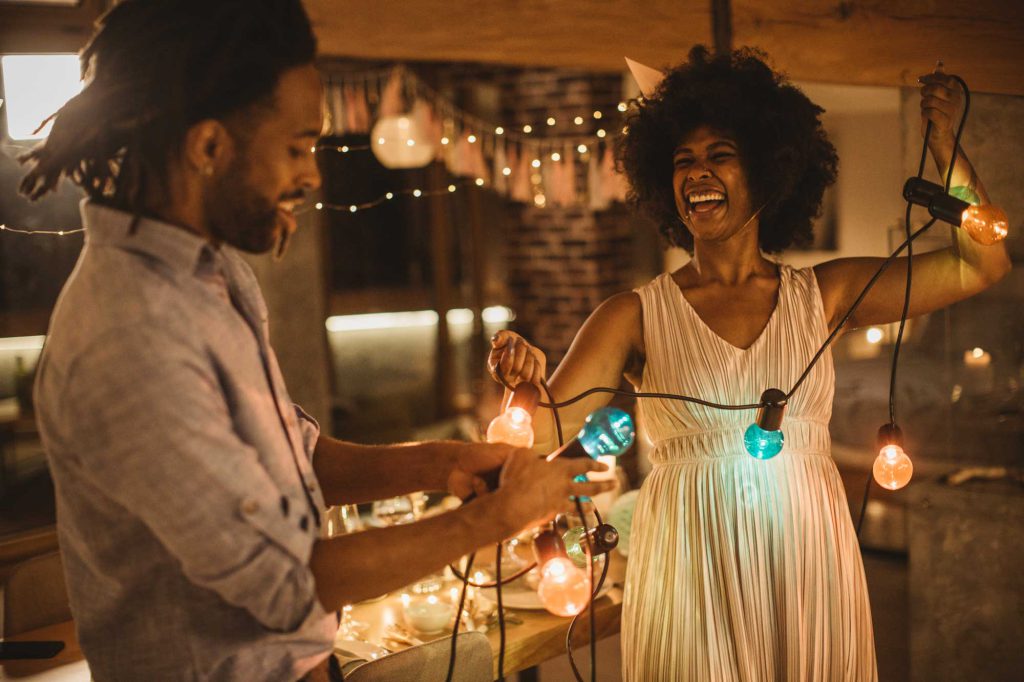 A couple laugh while hanging a string of lights inside near a set dining table.