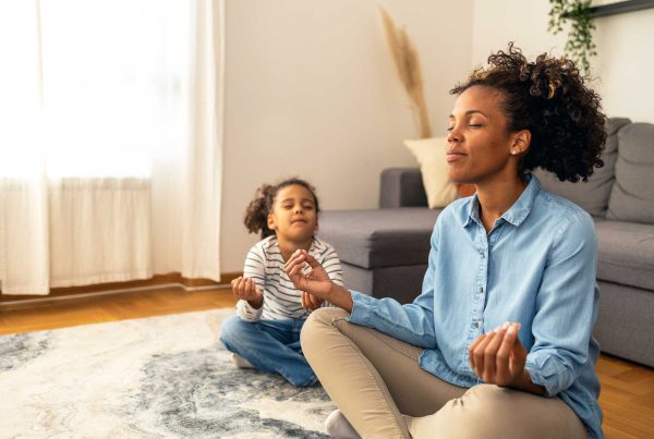 A mom and young daughter meditate in the easy pose on the rug in their clean and spacious living room.